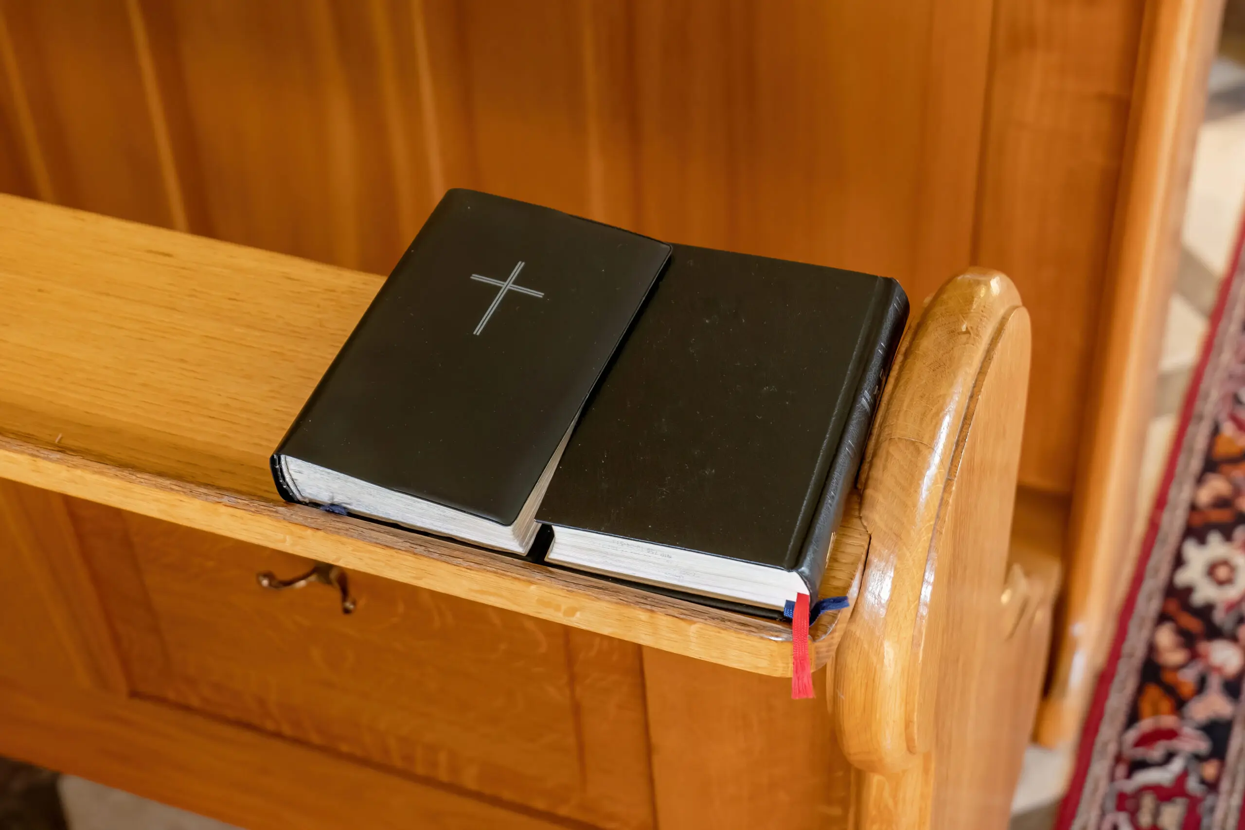 Black prayer book with a Christian cross symbol on it laying on a church pew bench, closeup detail shot, nobody. Bible, hymnal, gospel, small prayer books concept. Praying, prayers and faith, singing