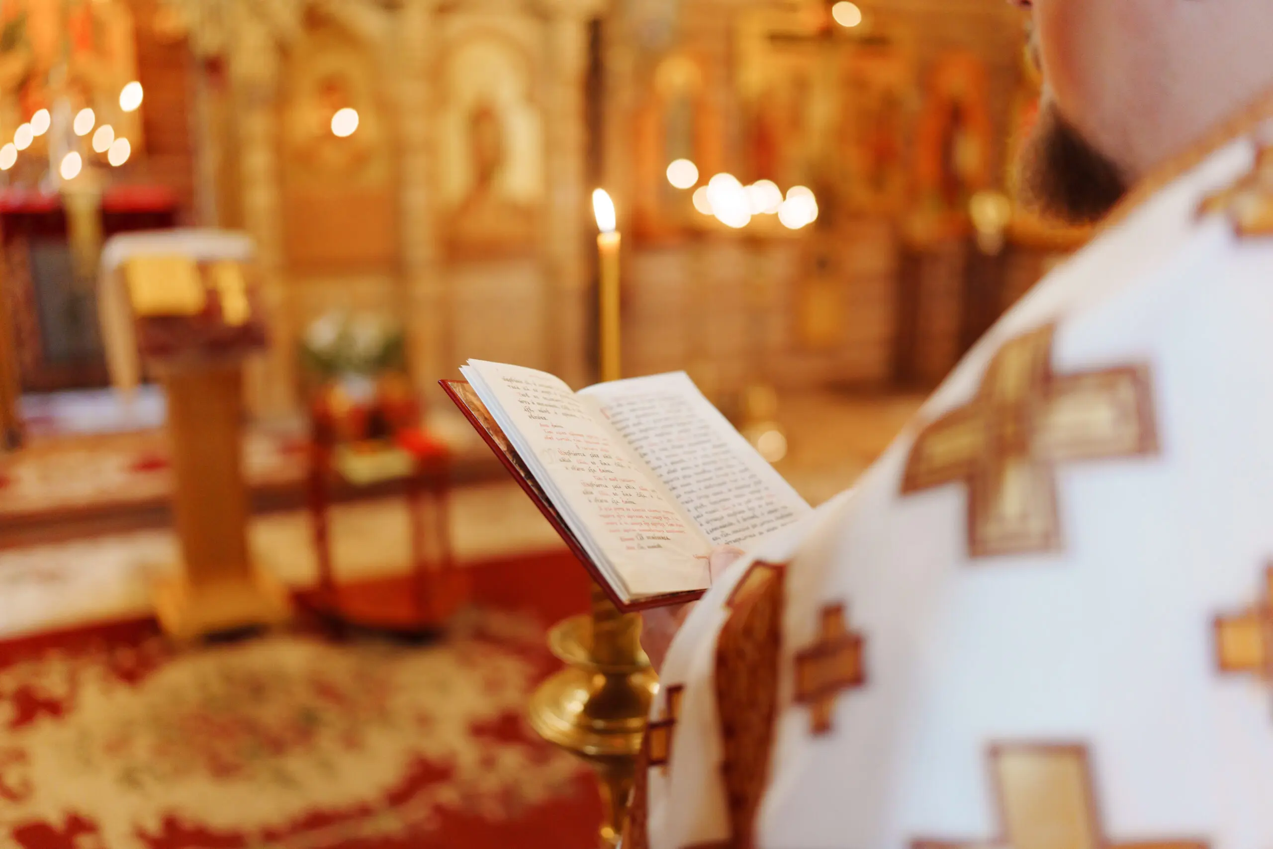 Inside a church, a priest stands holding an open book of scripture, focusing on the text. Candles illuminate the space, adding to the solemn atmosphere during the religious service.
